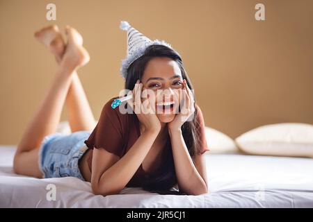 Felice giovane donna con cappello di compleanno sdraiato sul letto con il suo mento appoggiato sulle mani Foto Stock