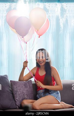 Giovane donna che tiene un mazzo di palloncini mentre si siede sul divano nel soggiorno Foto Stock