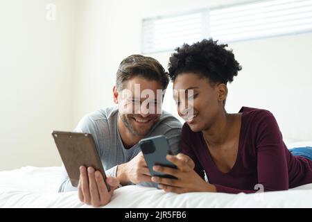 Immagine di una coppia felice e diversa utilizzando smartphone e tablet e sdraiata a letto Foto Stock