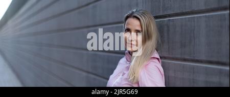 Giovane donna bionda in piedi davanti al muro di cemento. Fotografia ampia. Foto Stock