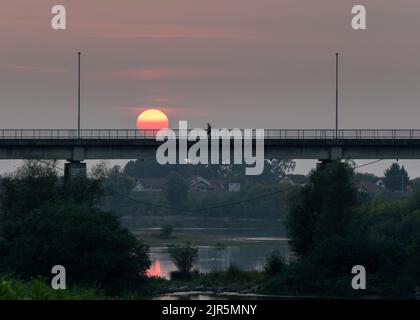 Un uomo cammina su un ponte sul fiume Sava tra Bosnia-Erzegovina e Croazia contro un cielo nuvoloso con un grande sole rosso. Foto Stock