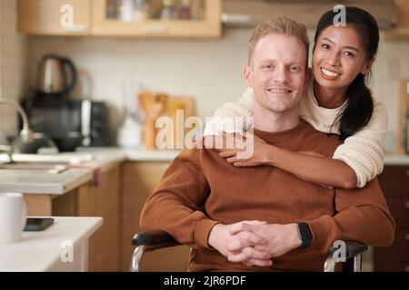 Giovane donna allegra che abbraccia un giovane seduto in sedia a rotelle Foto Stock