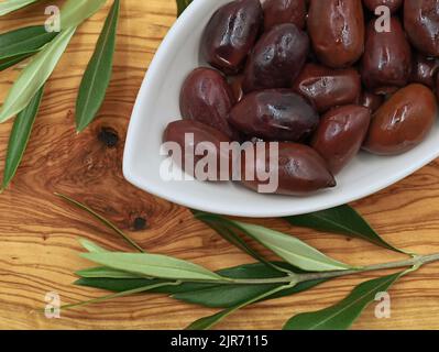 Olive Kalamata in ciotola bianca su tavola di legno d'oliva con ramo, vista dall'alto Foto Stock