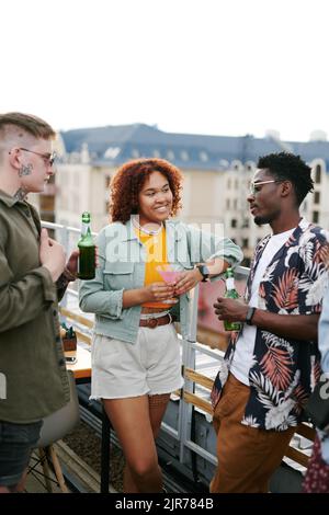 Due giovani coppie interculturali in un elegante casualwear sorseggiando un drink e chiacchierando sulla terrazza del caffè sul tetto durante una festa all'aperto Foto Stock