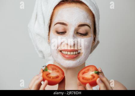 Ritratto di giovane donna caucasica con maschera di argilla bianca e asciugamano sul viso sorridente, mentre tiene due metà di pomodoro in mano. Isolato su sfondo grigio. Foto Stock