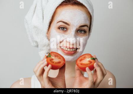 Ritratto di giovane donna caucasica con maschera di argilla bianca e asciugamano sul viso sorridente, mentre tiene due metà di pomodoro in mano. Isolato su sfondo grigio. Foto Stock