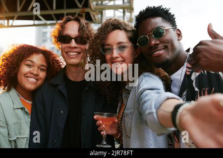 Due giovani e felici coppie interculturali in casualwear in posa per selfie alla festa mentre allegra ragazza con cocktail tenendo smartphone Foto Stock