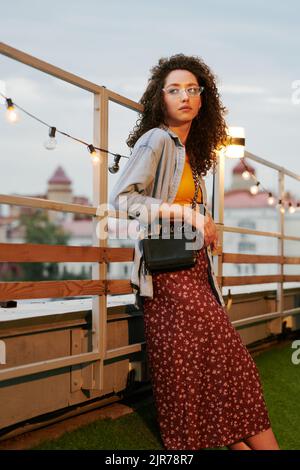 Giovane zoomer femminile in elegante casualwear con borsa in pelle nera sulla spalla in piedi contro l'intarsi decorata con illuminazione Foto Stock