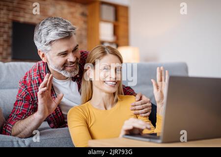 Felice sorridente marito caucasico di mezza età abbracciando moglie, facendo una mano nel laptop, facendo una chiamata in linea Foto Stock