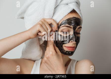 Bella giovane donna che rimuove la maschera cosmetica nera dal viso in asciugamano bianco su sfondo grigio. Bella signora che fa le procedure di cura della pelle in studio. Foto Stock