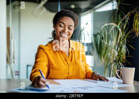 Felice lavoratrice di società nera che lavora con documenti, scrivendo e facendo documenti, seduto sul posto di lavoro in ufficio Foto Stock