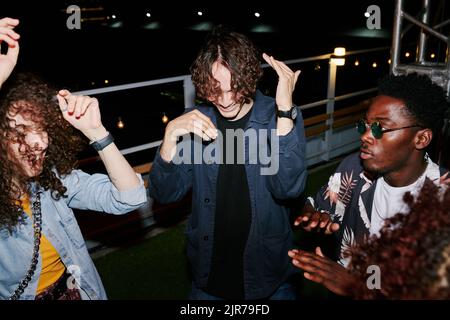 Due giovani coppie interculturali allegre in elegante casualwear ballare sul tetto di notte, mentre godersi la festa in caffè all'aperto Foto Stock