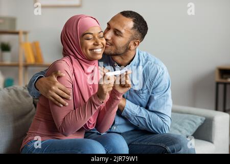 Sposi islamici neri felici che tengono il test positivo di gravidanza e baciano nel paese Foto Stock