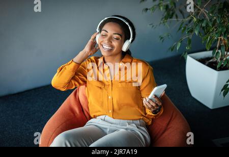 Felice imprenditore nero donna che indossa cuffie e godersi la musica, avendo pausa al lavoro seduto in beanbag sedia Foto Stock
