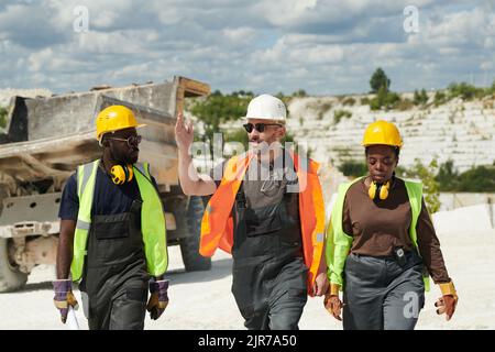 Caposquadra fiduciosa nel settore dell'abbigliamento da lavoro che spiega qualcosa ai suoi subordinati afro-americani maschi e femmine sulla strada per la fabbrica Foto Stock