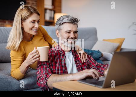 Felice moglie e marito caucasico maturo guardando il computer portatile, goditi una pausa caffè nel tempo libero nel soggiorno Foto Stock