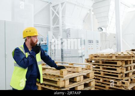 Giovane uomo sicuro ingegnere in guanti, abbigliamento da lavoro e casco di sicurezza parlando da walkie-talkie, mentre in piedi da una pila di pallet di legno Foto Stock
