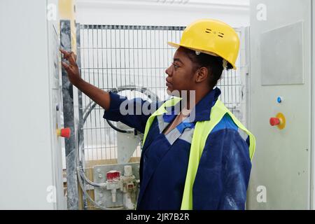 Vista laterale di una giovane donna nera in elmetto e uniforme davanti al pannello delle apparecchiature e premendo il pulsante di avviamento Foto Stock