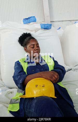 Donna afroamericana stanca con casco protettivo seduto contro un mucchio di enormi sacchi bianchi con materie prime in magazzino e napping Foto Stock