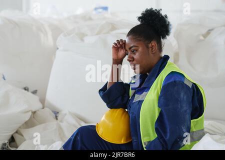 Giovane lavoratrice o lavoratrice stanca di magazzino seduta da un mucchio di enormi sacchi bianchi con materie prime e riposante dopo il lavoro Foto Stock