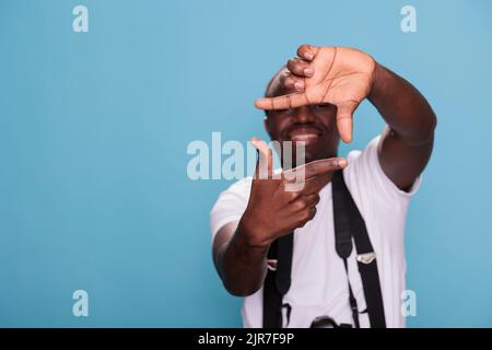 Fotografo professionista sicuro che fa gesti con le mani mentre ha una fotocamera moderna su sfondo blu. Ragazzo giovane con dispositivo DSLR gesturing scattare foto con le mani. Foto da studio Foto Stock
