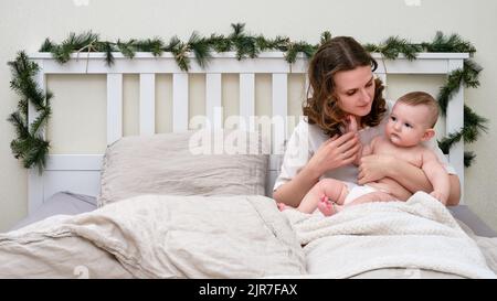 Donna triste con un bambino del bambino su un letto decorato per Natale la vigilia di Capodanno, famiglia nella camera da letto domestica Foto Stock