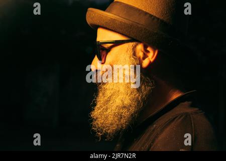 Un ritratto di un elegante uomo caucasico bearded in occhiali da sole e un cappello sullo sfondo nero Foto Stock