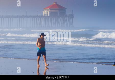 Uomo che cammina lungo la sabbia a Huntington Beach California indossando un cappello e un paio di pantaloncini la mattina presto con il molo nella nebbia di sfondo. Foto Stock