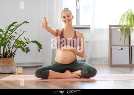 una donna incinta mostra i pollici seduti sul tappetino yoga Foto Stock