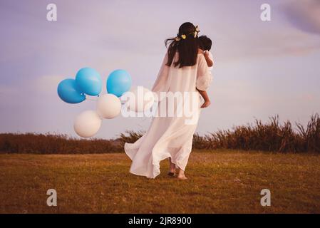 Tutto per lui. Ripresa da dietro di una giovane madre e suo figlio che cammina mentre tiene palloncini blu e bianchi all'aperto. Foto Stock