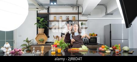 Le coppie asiatiche trascorrono del tempo insieme in cucina. Giovane donna in piatto di insalata di cottura grembiule mentre il suo ragazzo registra vlog video per il social blogger. Foto Stock