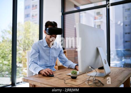 Portare il suo lavoro nel mondo virtuale. Un giovane uomo d'affari felice che indossa una cuffia per la realtà virtuale mentre lavora alla sua scrivania in ufficio. Foto Stock