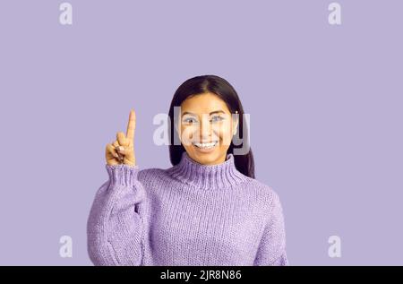 Bella giovane femmina con sorriso piacevole, fa gesto con indice dito, punta verso l'alto Foto Stock