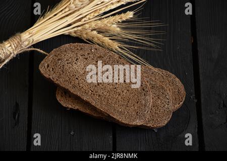 un pezzo di pane nero e spikelets secchi di grano su un tavolo di legno nero, pane sul tavolo Foto Stock