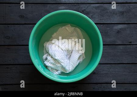 T-shirt bianca lavabile in acqua in una ciotola di plastica verde su un pavimento di legno nero, lavabile a mano in una ciotola Foto Stock