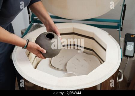 Primo piano delle mani mettere vaso nel forno, forno per tostare prodotti artigianali in argilla non cotta, ceramica non finita e arnese. Ceramica moderna Foto Stock