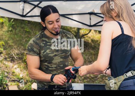 Muscular caucasian gun reseller in moro-modellato t-shirt mostrando le sue pistole al cliente femminile. Giorno in un campo di tiro. Riprese all'aperto. Foto di alta qualità Foto Stock