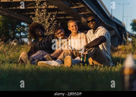 Felice gruppo variegato di giovani amici seduti insieme su una coperta sull'erba, bere birra, raccontare storie, ridere, e godersi la serata durante il loro viaggio in campeggio furgone. Foto di alta qualità Foto Stock