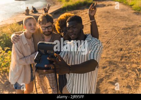 Gruppo multietnico di migliori amici creando ricordi scattando foto divertenti con un polaroide vintage, che si agghiacciano vicino al fiume all'ora del tramonto estivo . Foto di alta qualità Foto Stock