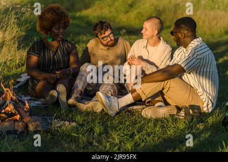 Positivo gruppo di amici diversi seduti su una coperta sull'erba vicino al fuoco, parlando e godendo il tramonto in natura. Escursione in campeggio con furgone selvaggio. Foto di alta qualità Foto Stock