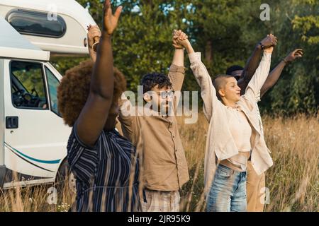 Gruppo di giovani felici migliori amici alzarsi e tenere le mani dopo aver raggiunto la destinazione del loro campeggio van viaggio su strada. Foto di alta qualità Foto Stock