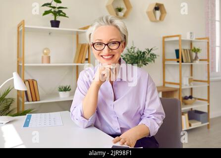Donna anziana felice con i capelli grigi sorridente guardando la macchina fotografica siede sul desktop in ufficio elegante Foto Stock