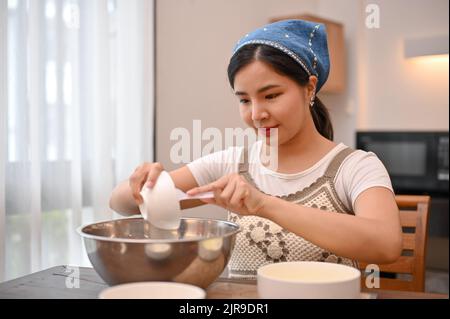 Attraente giovane donna asiatica fa un cupcake fatto in casa nella sua cucina, aggiungendo zucchero nel recipiente di miscelazione. Concetto di ricetta cupcake Foto Stock