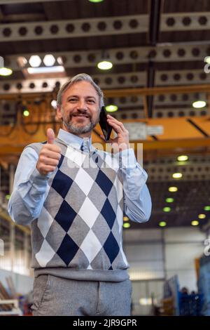 Vita Up Ritratto di sicuro, bello, grigio-capelli uomo d'affari o ingegnere in Industrial Interior. Uomo carismatico in piedi e utilizzando cellulare in Foto Stock