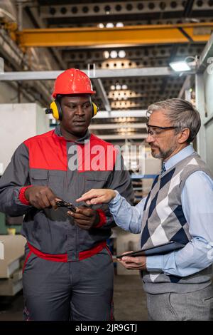 Lavoratori industriali multirazziali in piedi e parlare di processo di produzione. Misurazione del lavoratore nero con calibro a corsoio. Foto Stock