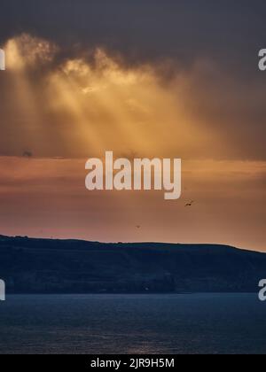 Le travi di un sole tramontato scoppiano attraverso pesanti nuvole sulla baia tra Whitby e Sandsend, raccogliendo gabbiani che si innalzano sulle scogliere e sul promontorio. Foto Stock