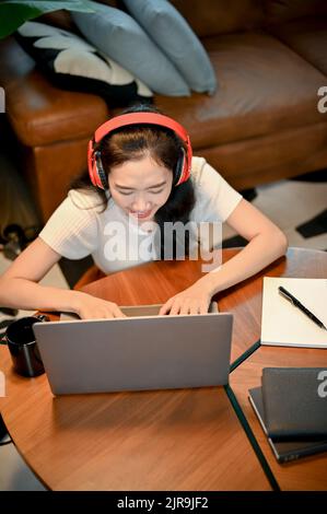 Sopra la vista, attraente giovane asiatica femminile in cuffie wireless si siede nel suo salotto e utilizza un computer portatile. Foto Stock