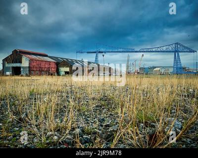 Vista a bassa angolazione di un rifiuto disseminato di erbacce, collegato a magazzini derelitti, corrosi con un cielo minaccioso e l'emblematico Transporter Bridge dietro. Foto Stock