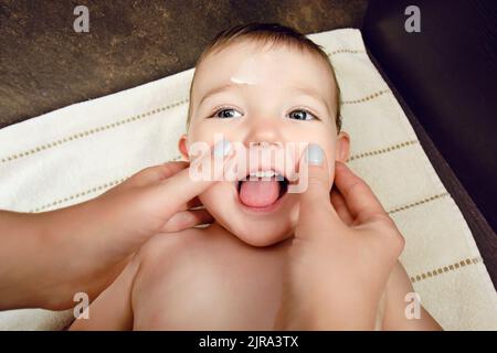 La madre spalma la crema sulla faccia di un bambino felice del toddler. La mamma spalma un unguento sulla pelle di un bambino sorridente. Bambino di un anno Foto Stock