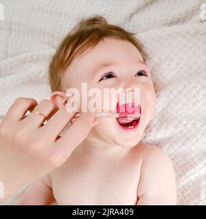 Le mani della madre stanno spazzolando i denti di un bambino felice del toddler su un letto domestico. La mamma spazzola la bocca del bambino con uno spazzolino. Bambino di un anno Foto Stock